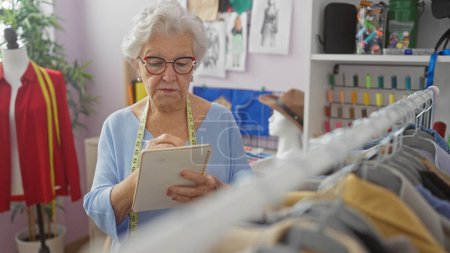 Téléchargez les photos : Une femme âgée avec des lunettes et un ruban à mesurer prend des notes dans une boutique de tailleurs colorés avec des mannequins et des tissus - en image libre de droit