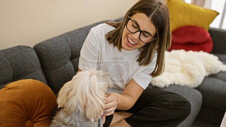Photo for A smiling young hispanic woman in glasses enjoys time with her fluffy dog in a cozy living room. - Royalty Free Image