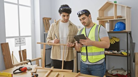 Foto de Dos hombres colaborando en un brillante taller de carpintería con herramientas, discutiendo los planes de proyecto en una tableta. - Imagen libre de derechos