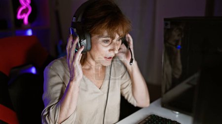 A mature woman wearing headphones focused on a computer screen in a dimly-lit gaming room at night.