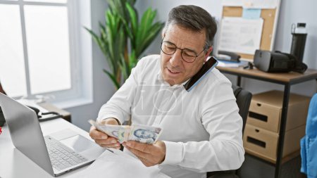 Un hombre maduro multitarea en una oficina, analizando dólares singapurenses mientras conversa en un teléfono inteligente.