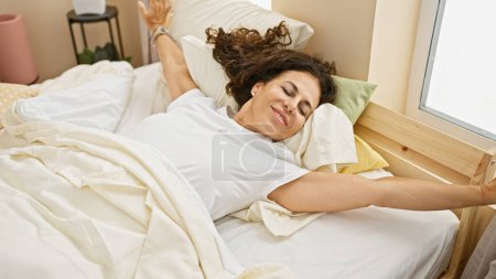 Foto de Una mujer hispana de mediana edad con el pelo rizado disfruta de un relajante estiramiento matutino en la comodidad de su dormitorio en casa. - Imagen libre de derechos