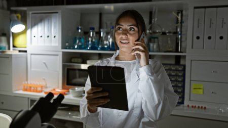 Photo for Hispanic woman in lab coat multitasking with tablet and phone in a laboratory setting, looking focused and engaged. - Royalty Free Image
