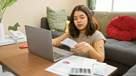 Hispanic woman working remotely with laptop in home interior examining paper