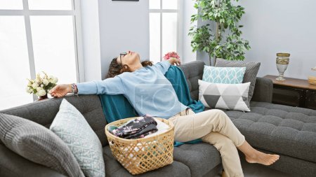 Foto de Relajada mujer de mediana edad reclinada cómodamente en un sofá gris en una acogedora y bien decorada sala de estar, gafas puestas, exhibiendo soledad y tranquilidad. - Imagen libre de derechos