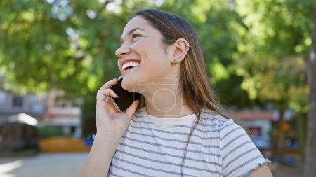 Téléchargez les photos : Une jeune femme joyeuse parlant sur un smartphone dans un parc ensoleillé avec une végétation luxuriante en arrière-plan - en image libre de droit