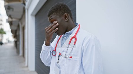 A tired black man in a doctor's coat with a stethoscope stands stressed on a city street.