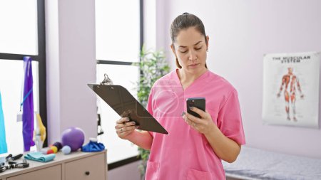 Gesundheitshelferin in rosa Peelings benutzt Smartphone, während sie Klemmbrett im Therapieraum einer Klinik hält