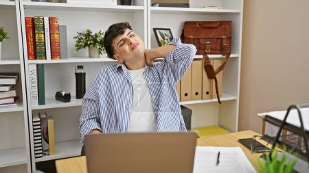 Téléchargez les photos : Un jeune homme s'étirant à son bureau organisé montrant de la fatigue après avoir travaillé sur un ordinateur portable. - en image libre de droit