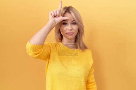 Photo for Young caucasian woman wearing yellow sweater making fun of people with fingers on forehead doing loser gesture mocking and insulting. - Royalty Free Image