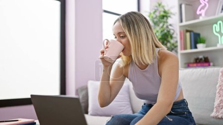 Téléchargez les photos : Jeune femme blonde utilisant un ordinateur portable boire du café à la maison - en image libre de droit
