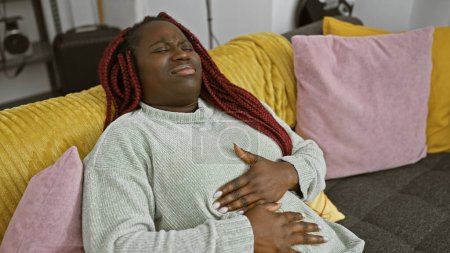 A black woman with braids expressing stomach pain while sitting on a couch indoors.