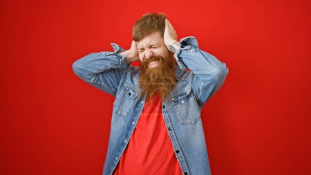 Téléchargez les photos : Stressé jeune rousse homme fatigué couvrant les oreilles sur l'anxiété bruyante, isolé sur fond rouge, exprimant problème de céphalée émotionnelle - en image libre de droit