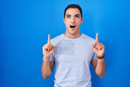 Foto de Young hispanic man standing over blue background amazed and surprised looking up and pointing with fingers and raised arms. - Imagen libre de derechos