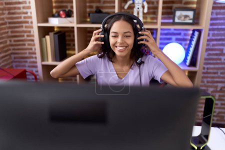 Téléchargez les photos : Jeune femme afro-américaine streamer souriant confiant assis sur la table à la salle de jeux - en image libre de droit