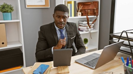 Photo for A professional african american man in a suit seated thoughtfully at his office desk with a laptop and tablet. - Royalty Free Image