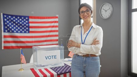 Foto de Mujer madura hispana con los brazos cruzados sonriendo con confianza en un centro de votación de Estados Unidos con urnas y bandera - Imagen libre de derechos