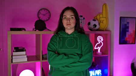 Confident young woman standing arms crossed in a neon-lit gaming room looking serious