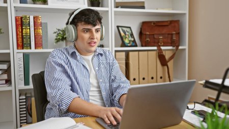 Foto de Joven con auriculares usando portátil en un moderno entorno de oficina, rodeado de libros y plantas. - Imagen libre de derechos