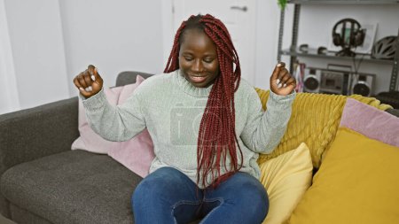 Foto de Una mujer africana alegre con trenzas rojas celebrando en el interior en una acogedora sala de estar con cojines de colores. - Imagen libre de derechos