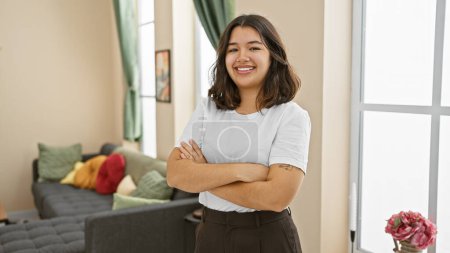Confident young hispanic woman posing indoors with arms crossed in a well-lit living room, exuding positivity and style.