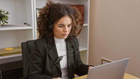 A young hispanic woman with curly hair works attentively at her laptop in an office setting, wearing headphones and a blazer.