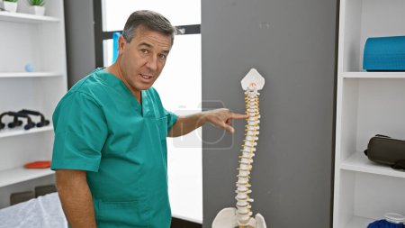 Confident male doctor in scrubs pointing at a spine model in a bright rehabilitation clinic.
