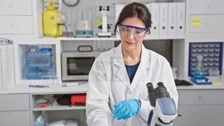 Foto de Científica madura en laboratorio con microscopio, guantes, gafas de seguridad, bata blanca y equipo en interiores. - Imagen libre de derechos