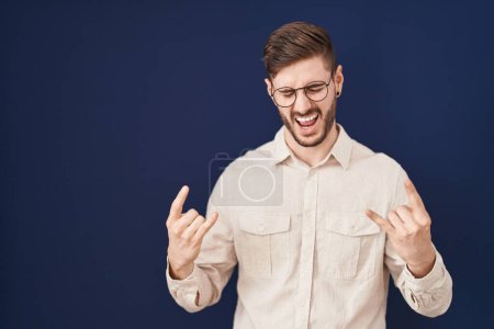 Téléchargez les photos : Homme hispanique avec barbe debout sur fond bleu criant avec une expression folle faire symbole rock avec les mains en l'air. star de la musique. concept lourd. - en image libre de droit