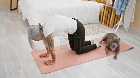 Téléchargez les photos : Femme sportive d'âge moyen, aux cheveux gris et en forme, entraîne le yoga avec son chien bien-aimé, lui étirant le dos dans l'ambiance relaxante de sa chambre - en image libre de droit