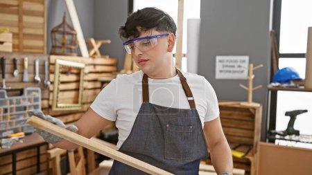 Téléchargez les photos : Jeune homme examine la planche de bois dans un atelier de menuiserie bien équipé. - en image libre de droit