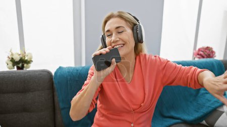 Foto de Una mujer madura alegre disfruta de la música en los auriculares en casa, cantando en su teléfono inteligente como un micrófono improvisado. - Imagen libre de derechos