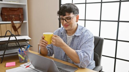 Photo for A young man enjoys a break with coffee at his modern office desk, displaying casual professionalism and comfort. - Royalty Free Image