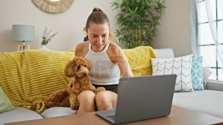 Téléchargez les photos : Jeune femme caucasienne avec chien souriant à l'aide d'un ordinateur portable assis sur le canapé à la maison - en image libre de droit