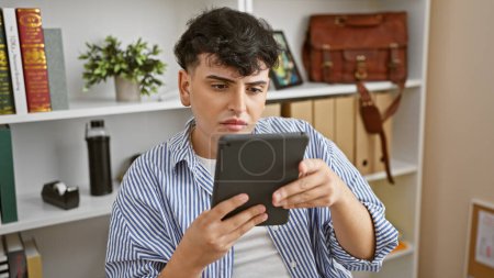 Téléchargez les photos : Un jeune homme en chemise rayée, concentré sur une tablette dans un cadre de bureau moderne, exprimant concentration et détermination. - en image libre de droit