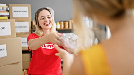 Téléchargez les photos : Jeune femme blonde bénévole souriant confiant donnant sandwich à la personne au centre de charité - en image libre de droit