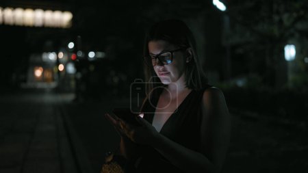 Photo for At night, beautiful hispanic woman wearing glasses holds a deep conversation over text, engrossed in her smartphone on kyoto street, amid the city's twinkling lights. - Royalty Free Image
