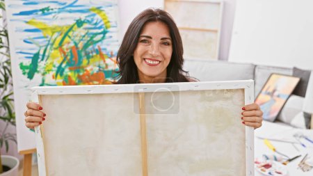 Photo for Portrait of a smiling brunette woman holding a blank canvas in an art studio filled with paintings and art supplies. - Royalty Free Image