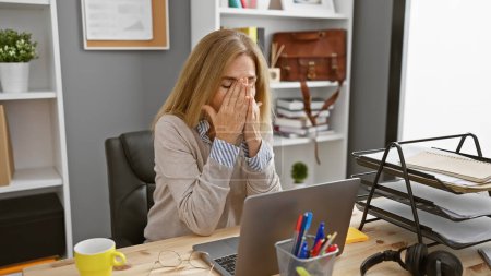 Téléchargez les photos : Stressé femme d'âge moyen frottant les yeux au bureau avec ordinateur portable - en image libre de droit
