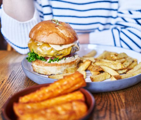 Woman enjoying a gourmet burger with fries and sweet potato fries in a casual dining setting.