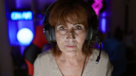 A mature woman wearing headphones focuses intently in a neon-lit gaming room at night.