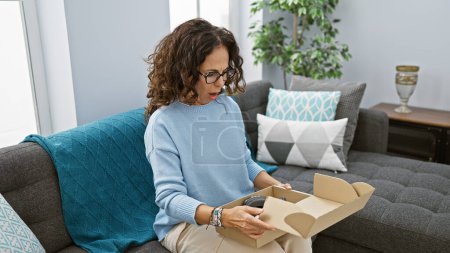 Photo for Surprised woman unpacking headphones in cozy living room, expressing delight and curiosity. - Royalty Free Image