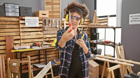Téléchargez les photos : Femme hispanique aux cheveux bouclés portant des lunettes de sécurité dans un atelier de menuiserie tenant des outils - en image libre de droit