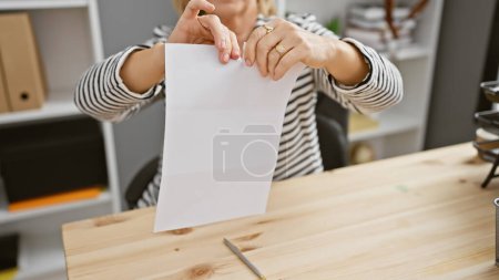 A middle-aged woman tears a white paper in an office setting, displaying a mix of frustration and determination.