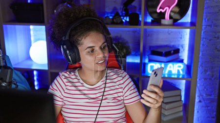 A young hispanic woman with curly hair enjoys using her smartphone in a modern gaming room at night, exuding beauty and happiness.