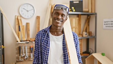 Foto de Un hombre africano sonriente en un taller de carpintería con gafas de seguridad y ropa casual. - Imagen libre de derechos