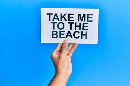 Hand of caucasian man holding paper with take me to the beach message over isolated blue background