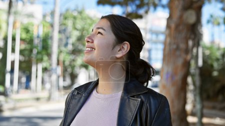 Foto de Mujer joven hispana sonriente con atuendo casual disfrutando del aire libre en un parque de la ciudad con árboles y luz del día. - Imagen libre de derechos