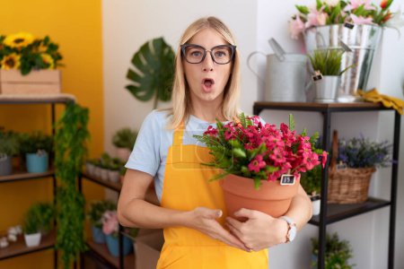Photo for Young caucasian woman working at florist shop holding plant scared and amazed with open mouth for surprise, disbelief face - Royalty Free Image