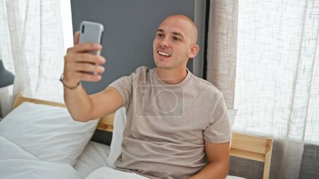 Téléchargez les photos : Jeune homme hispanique faire selfie par smartphone assis sur le lit dans la chambre - en image libre de droit